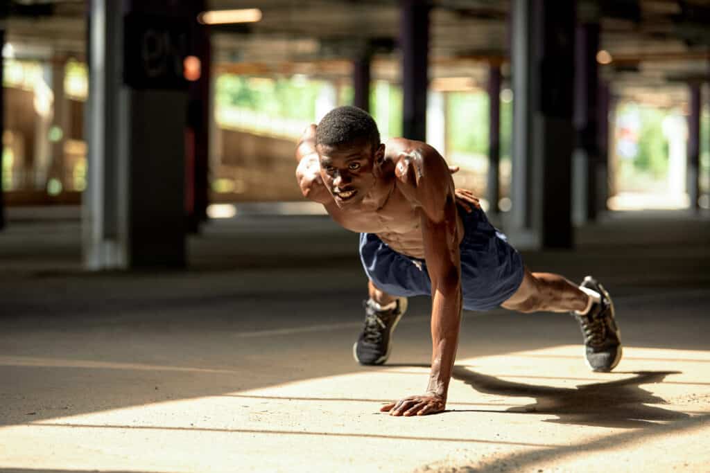 Side view of muscular black man in sportswear doing push ups during workout on street of modern downtown with urban background.